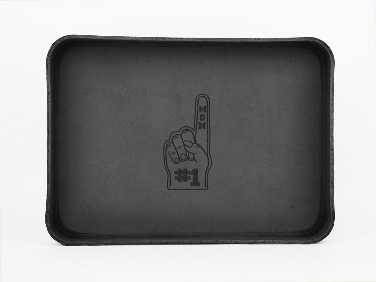 A black rectangular tray with a line drawing of a foam finger in the center. The finger points up and reads MOM on the finger and #1 on the palm, symbolizing Number 1 Mom.