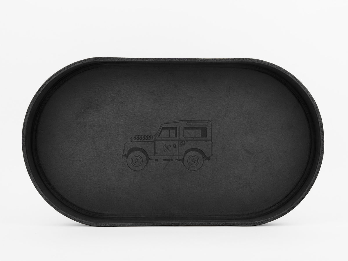 A black, oval-shaped tray with an outline drawing of a classic SUV, similar to a Land Rover, engraved in the center. The background is plain white.