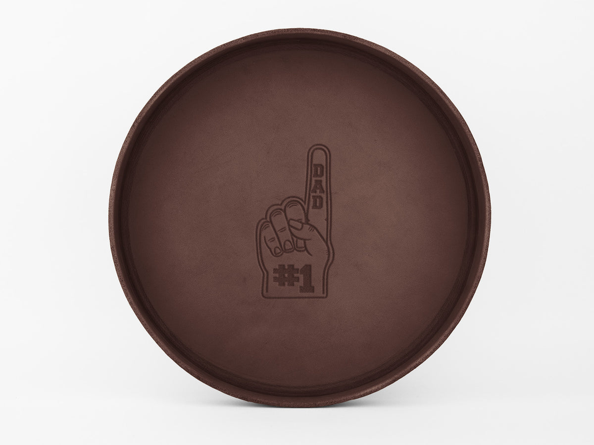 A round brown tray with an embossed image of a foam finger that reads “DAD” on the finger and “#1” on the palm, centered on the tray’s surface.
