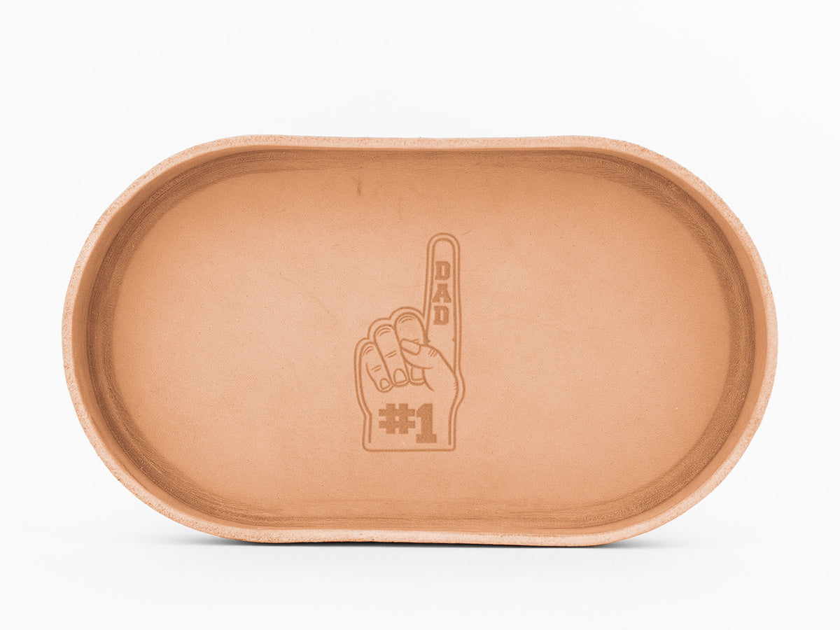 Oval-shaped, natural colored leather tray with embossed image of a hand holding up a foam finger. The finger has the word DAD on it, and below the hand is the text #1, resembling a foam finger sign.