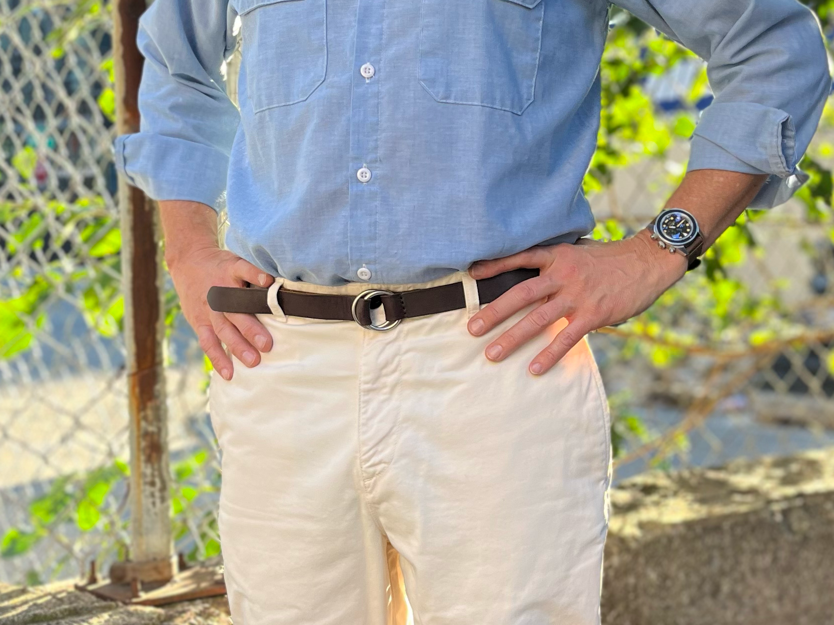 A person stands outdoors with hands on hips, wearing a light blue button-up shirt, cream-colored pants, a dark brown belt, and a wristwatch. Sunlight and a chain-link fence are visible in the background.