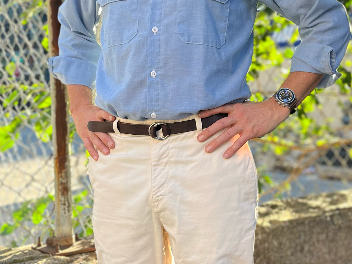 A person stands outdoors with hands on hips, wearing a light blue button-up shirt, cream-colored pants, a brown belt, and a wristwatch. Sunlight and a chain-link fence are visible in the background.