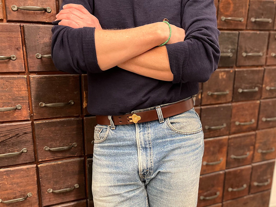 Person standing with arms crossed, wearing a navy sweater, light blue jeans, and a brown belt with a gold buckle, in front of a wall of vintage wooden drawers with metal handles.