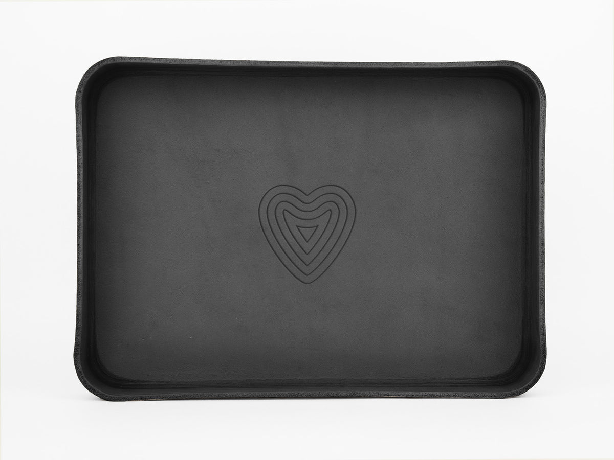 A black rectangular tray with rounded edges, featuring a stylized heart design embossed in the center, set against a plain white background.