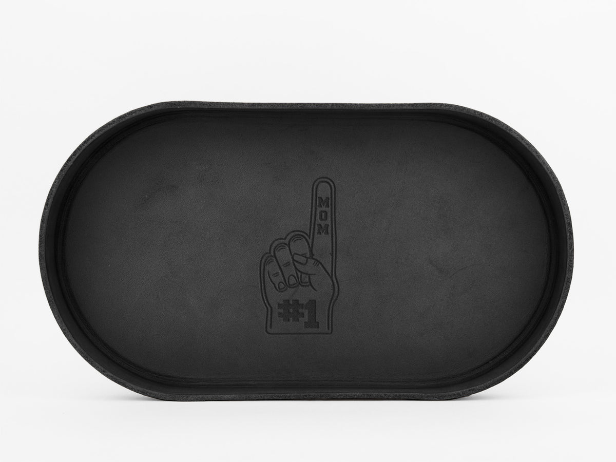 A black oval tray with an embossed design of a foam finger hand. The finger points upward and reads MOM with #1 at the base, all in black on the trays surface.