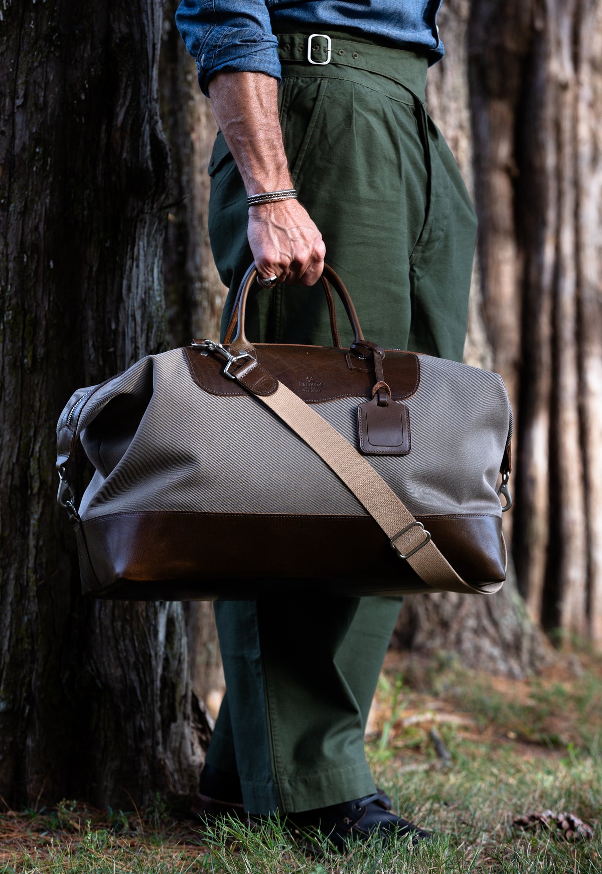 Person holding a beige duffel bag with olive leather accents in a forest setting