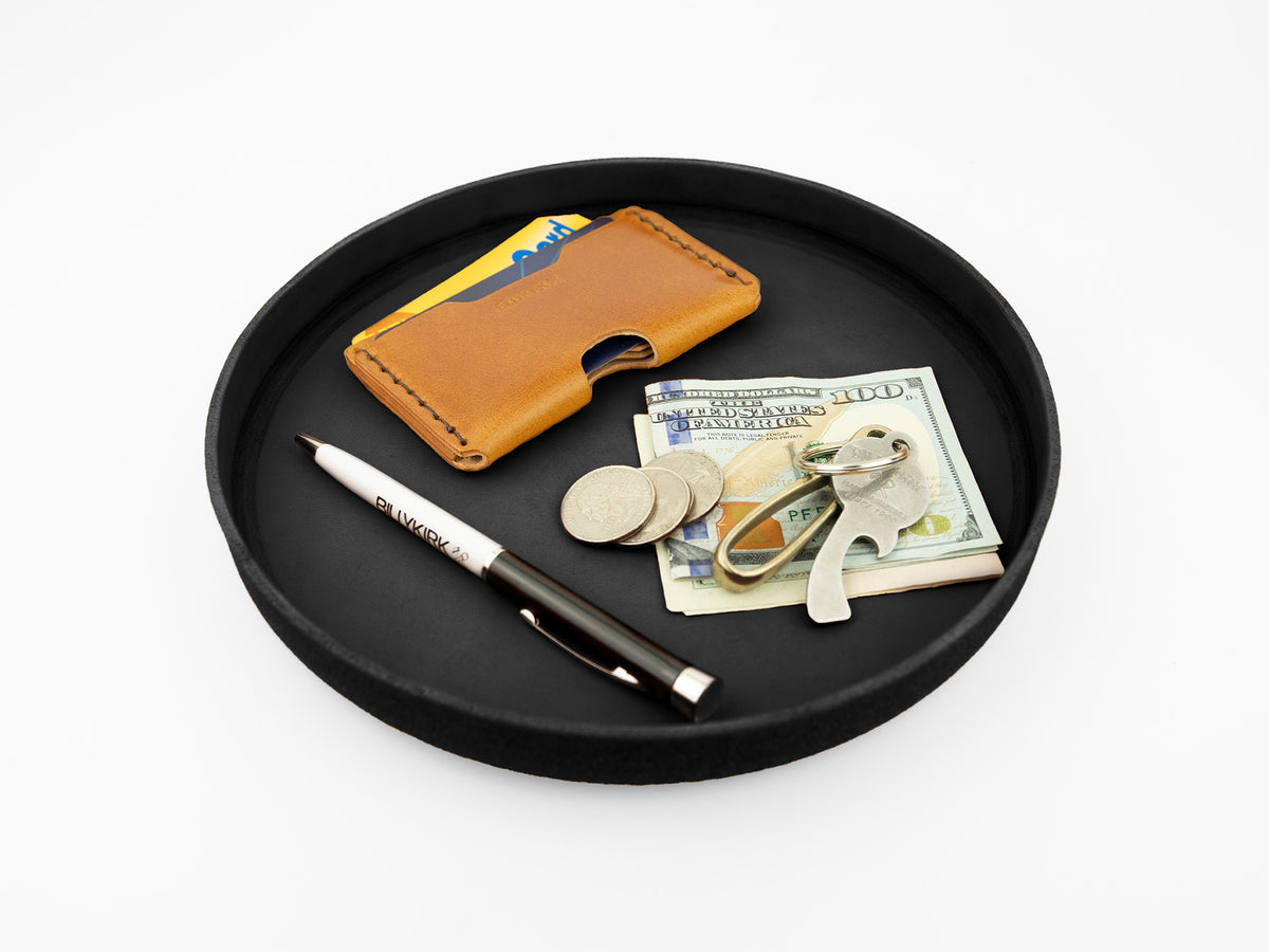 A black tray holds a brown leather card holder, a few credit cards, a pen, several coins, a set of keys, and folded U.S. dollar bills on a white background.