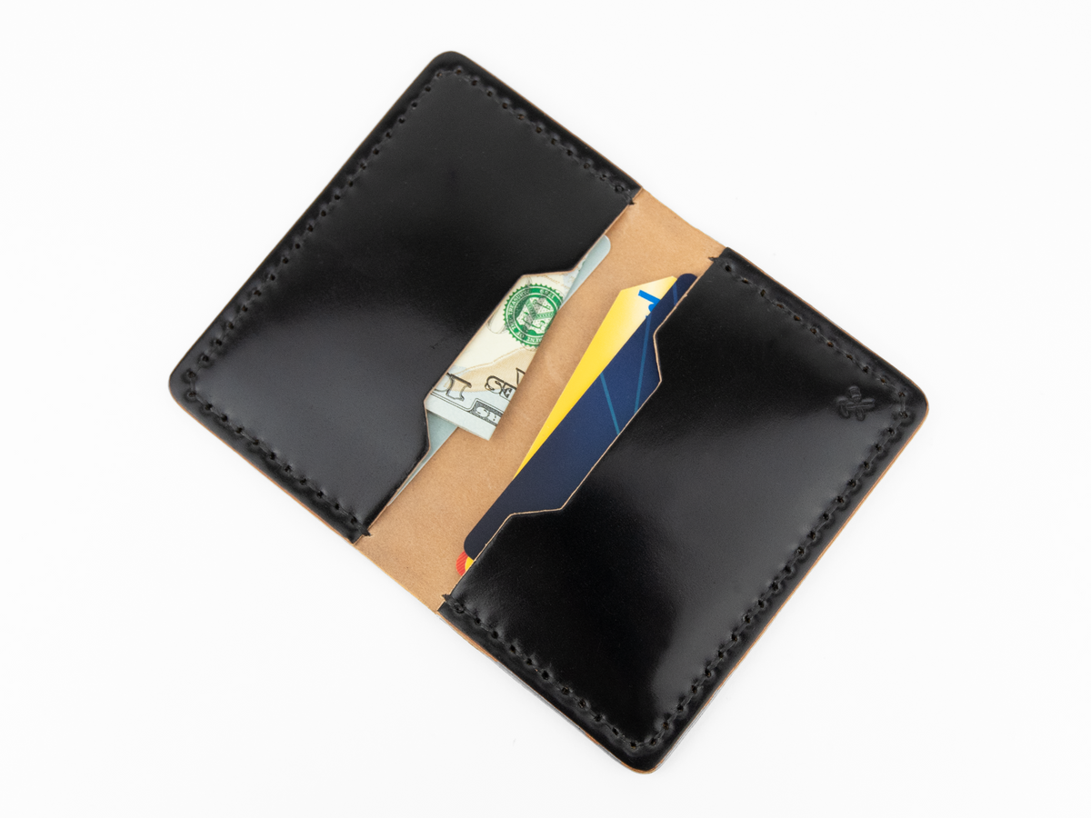 A black cordovan leather wallet open to reveal paper currency and two credit cards inside, photographed on a white background.