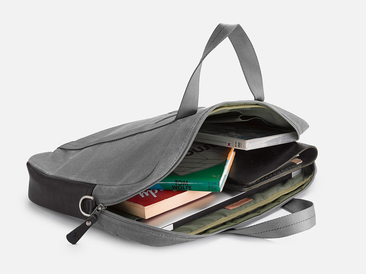 A gray fabric briefcase lies open, revealing books, a notebook, and a tablet inside. The bag has two handles and is placed on a white background.