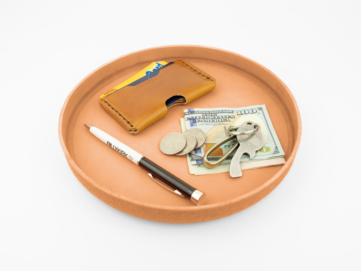 A round brown tray holds a tan leather card holder with credit cards, a pile of coins, a stack of cash, keys, and two pens, all arranged neatly on a white background.