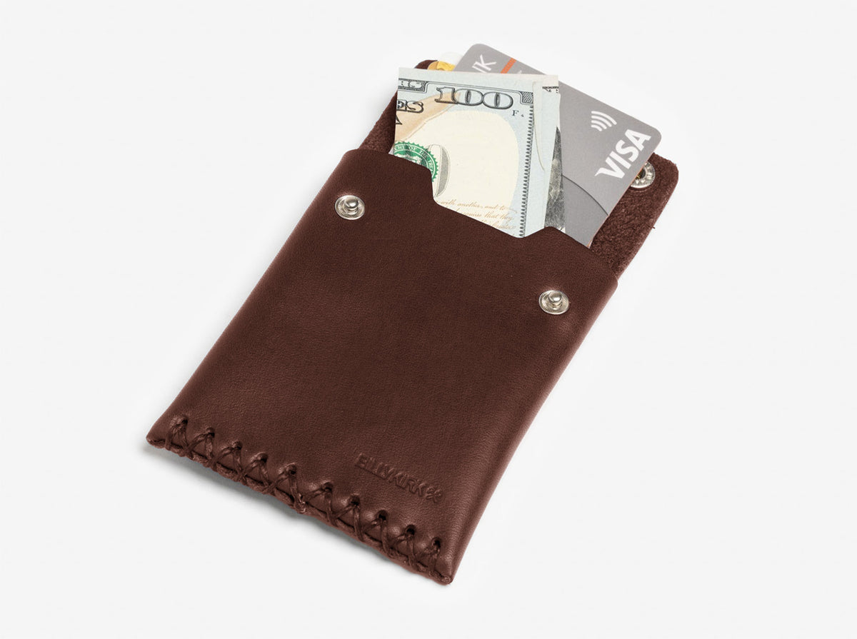 A brown leather card holder with visible stitching contains a folded U.S. hundred-dollar bill and two credit cards, one labeled Visa, partially sticking out from the top.