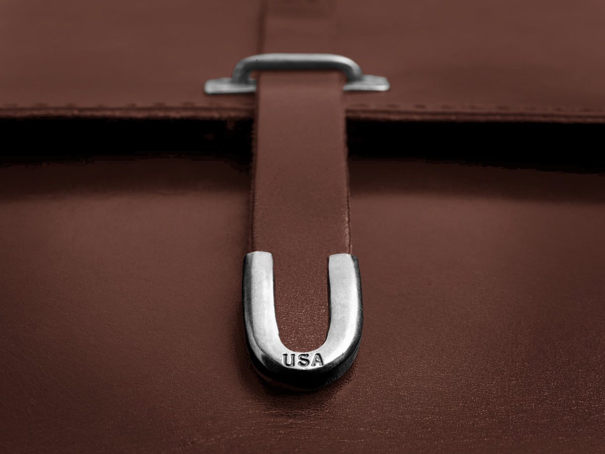 Close-up of a brown leather bag with a metal U-shaped clasp engraved with the letters USA. The clasp secures a leather strap on the bag.