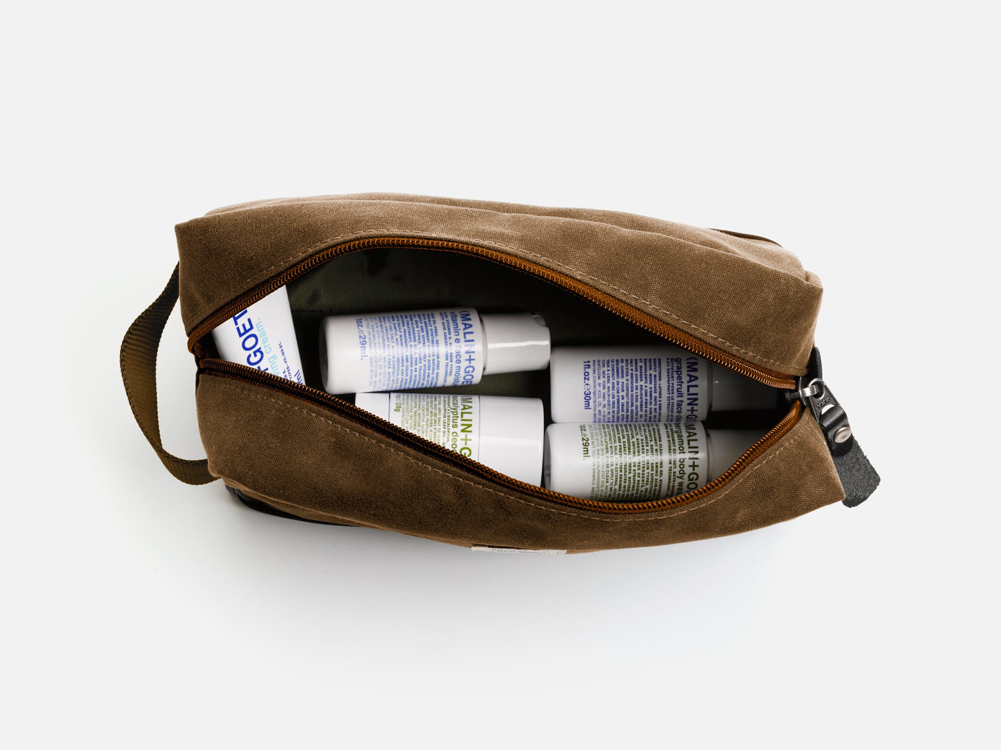 A brush brown waxed canvas toiletry bag unzipped to reveal several Malin+Goetz branded bottles and containers inside, arranged neatly against a white background.