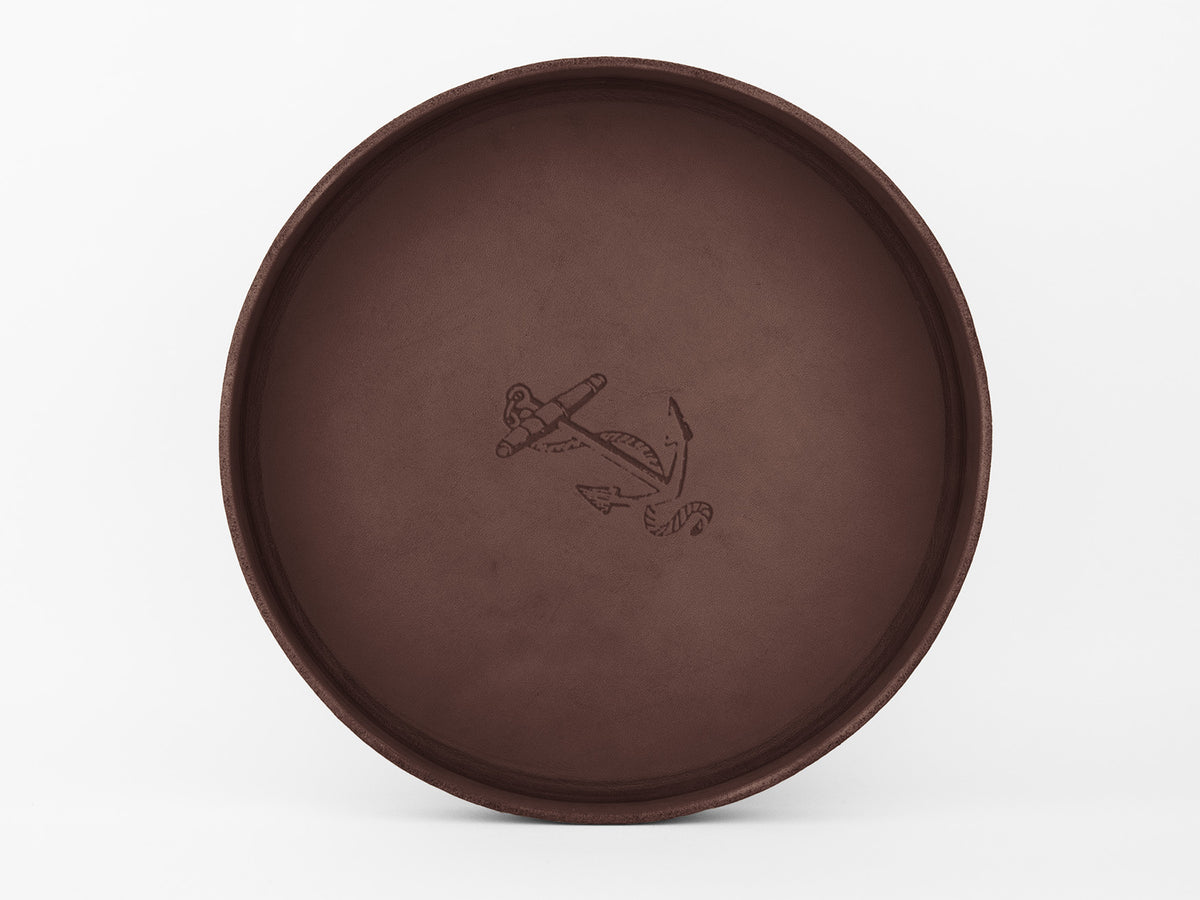 	
A round, brown leather tray with a simple anchor and rope design engraved in the center, set against a plain white background.