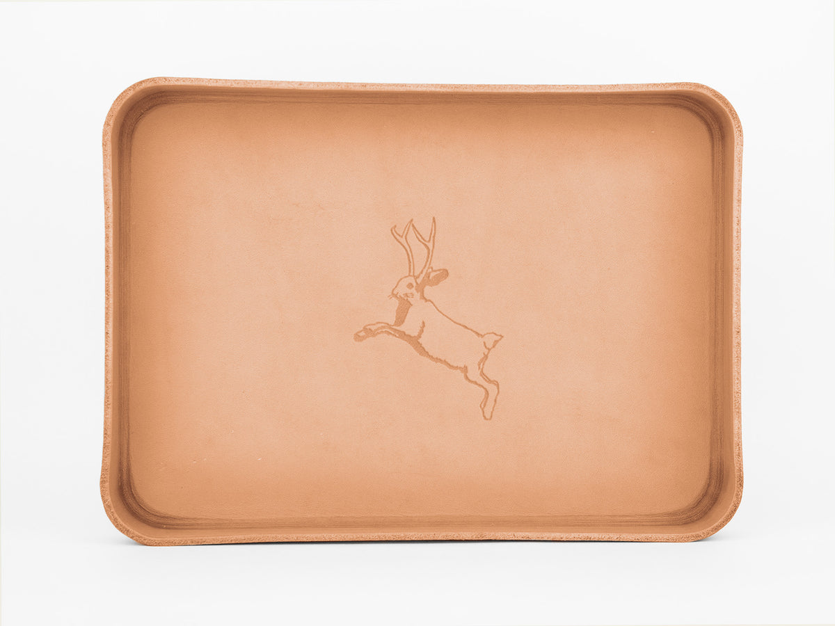 A rectangular tan leather tray with raised edges, featuring an engraved outline of a leaping jackalope (a rabbit with antlers) in the center. The background is plain white.