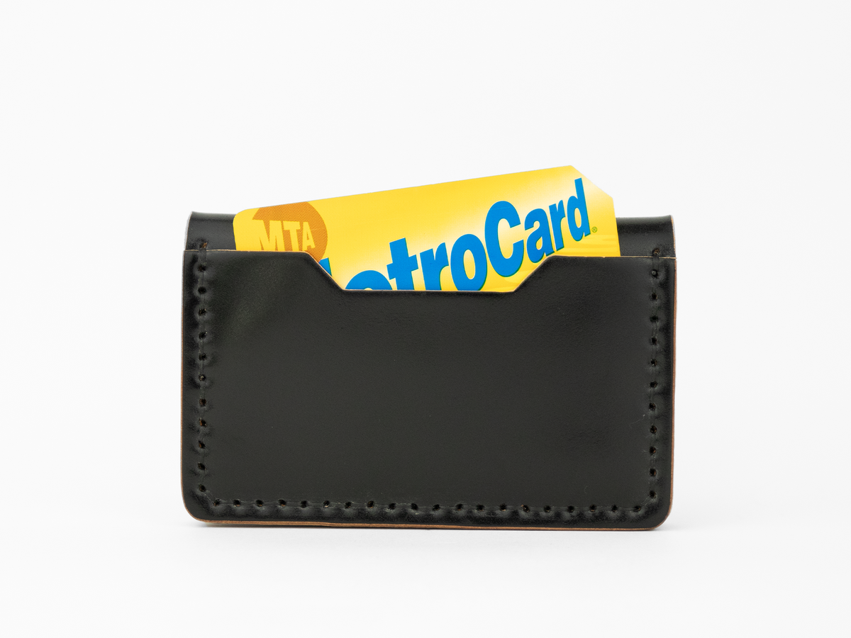 A black cordovan leather card holder with visible stitching holds a yellow and blue MTA MetroCard, partially sticking out from the top.