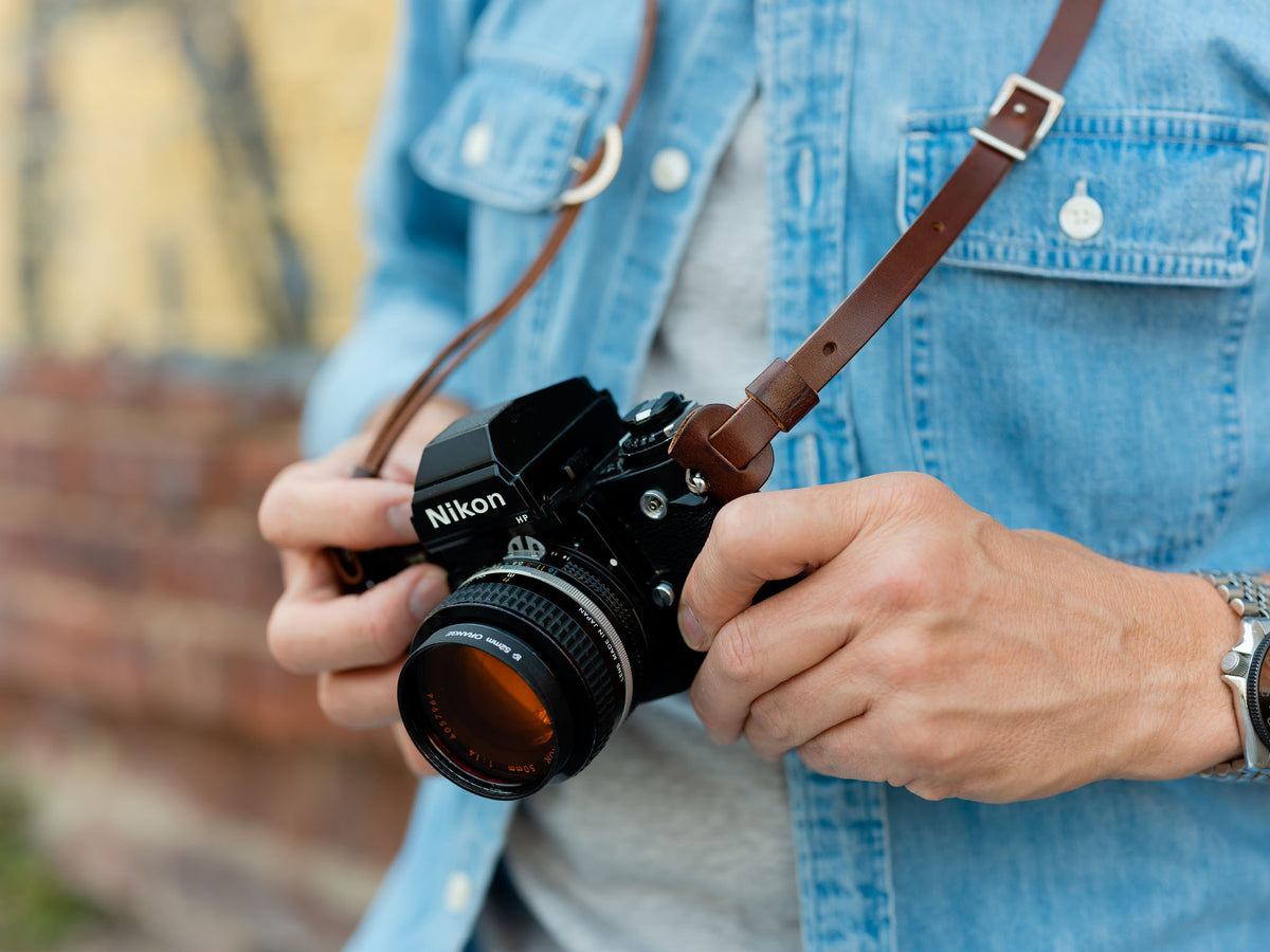 A person wearing a denim shirt holds a Nikon film camera with both hands, the camera hanging from a brown leather strap around their neck.