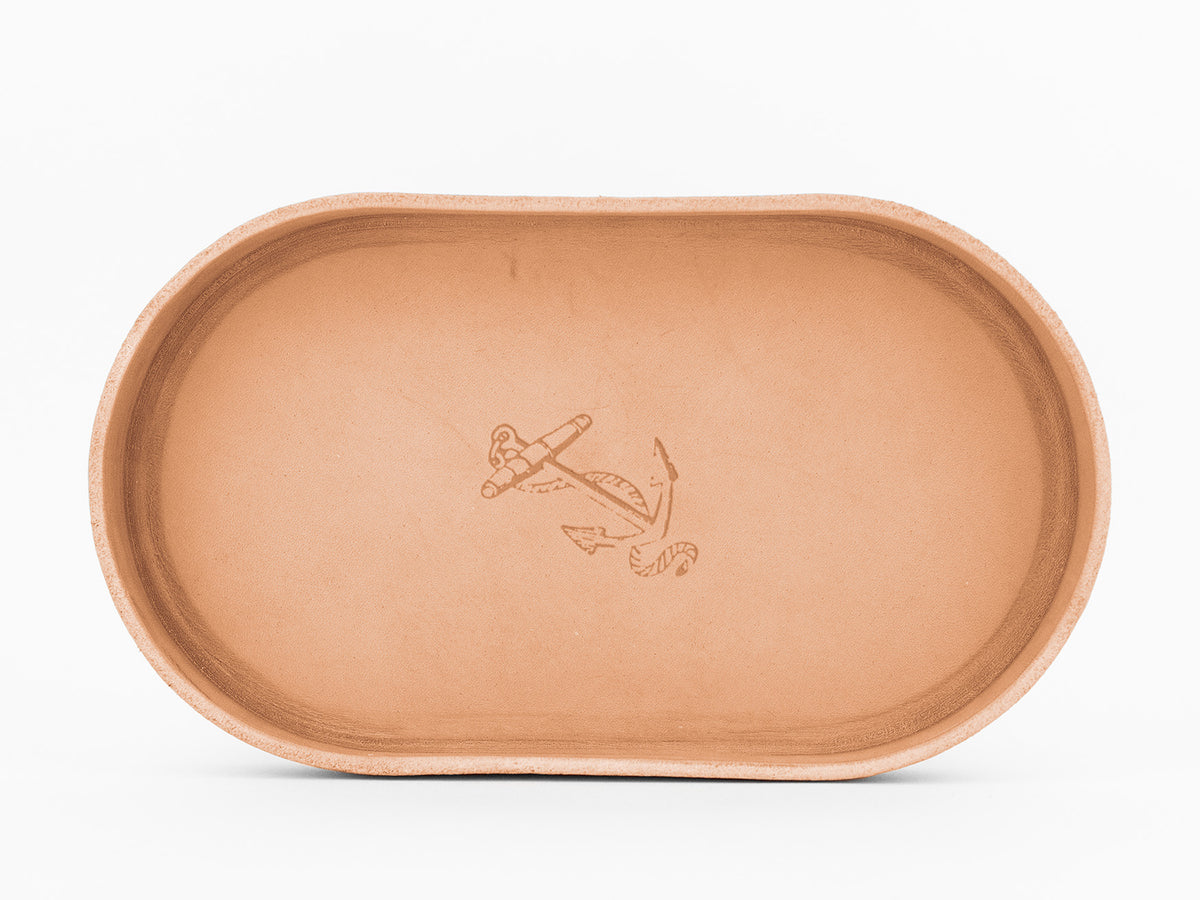 A natural, oval-shaped leather tray with a simple anchor and rope design engraved in the center. The tray has smooth edges and a minimalist style.