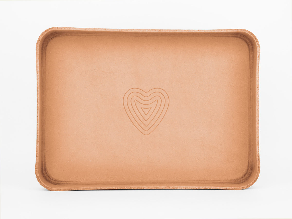 A rectangular tan leather tray with raised edges and a subtle, concentric heart design embossed in the center, set against a white background.