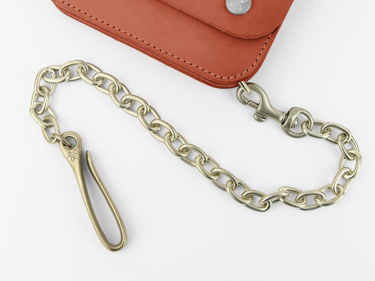 A brass chain with a hook and clasp is attached to a brown leather wallet with visible stitching and metal rivets. The chain is coiled in a loose curve on a white surface.