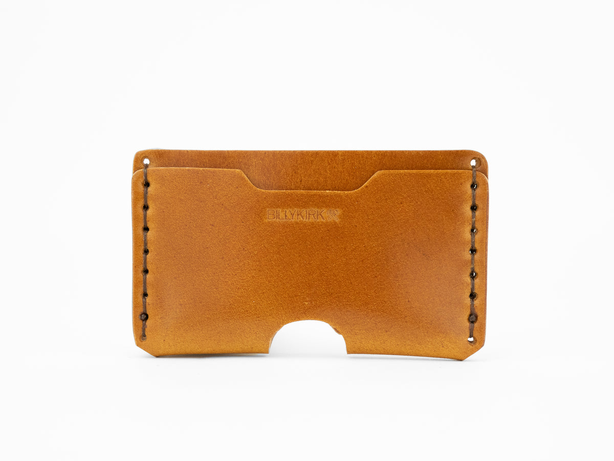 A tan cordovan leather wallet with visible stitching along the sides, an open slot on top, and a semicircular cutout at the bottom center. The word BILLYKIRK is embossed on the front.