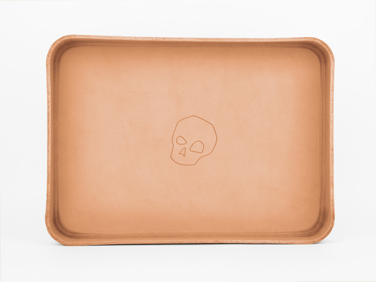 A rectangular, tan leather tray with rounded edges. The tray features a simple, embossed skull design in the center. The background is plain white.