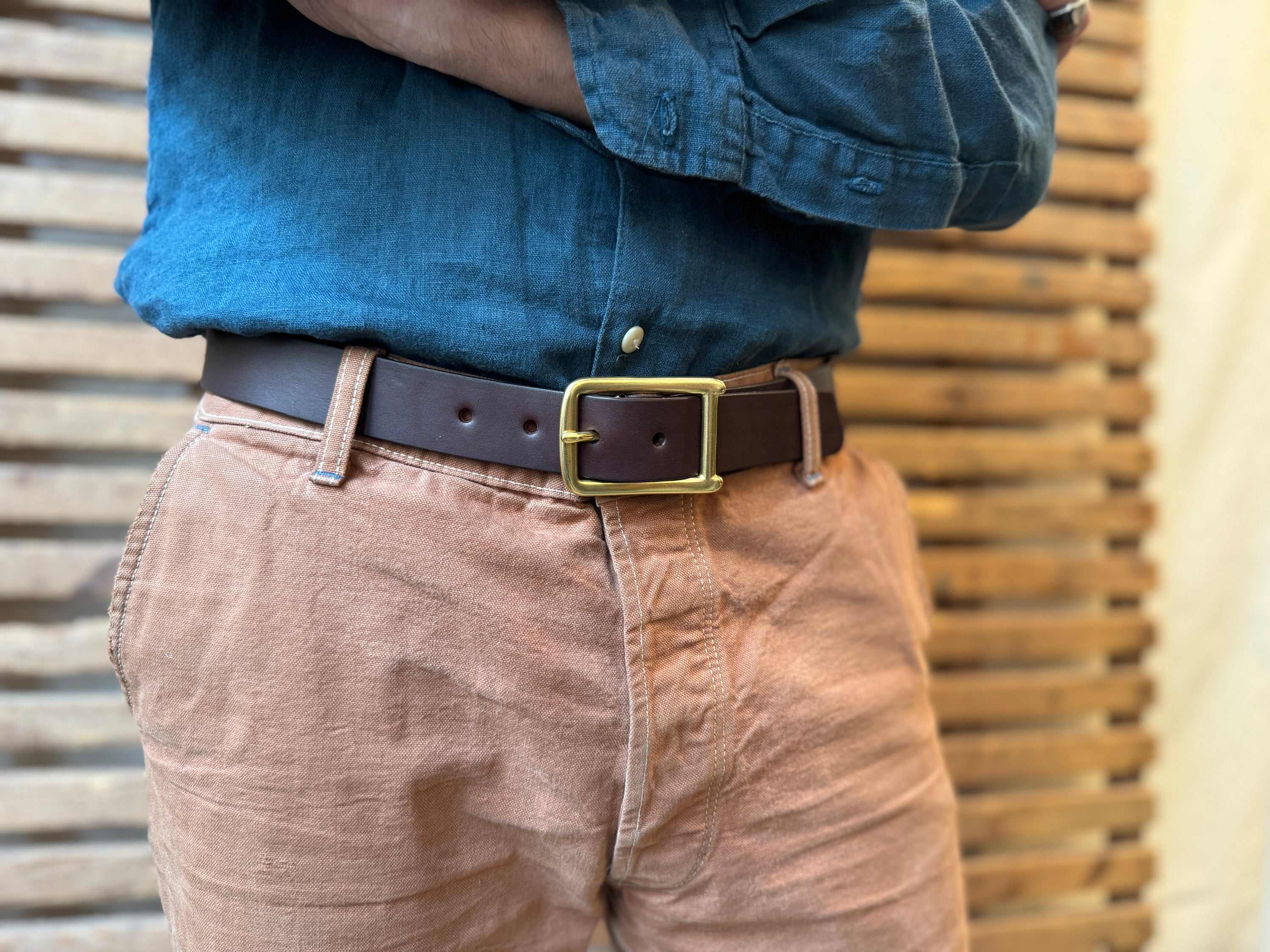 A person wearing a blue denim shirt tucked into light brown pants with a brown leather belt and gold buckle, standing in front of a wooden slat background.