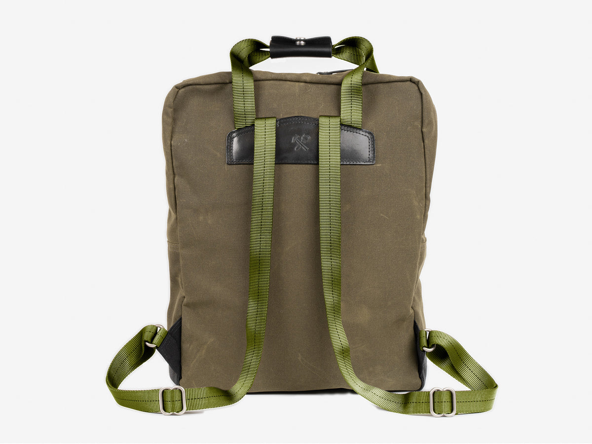 A olive waxed canvas backpack shown from the back, featuring adjustable shoulder straps, a top handle, and a rectangular black leather patch near the top center. The background is plain white.