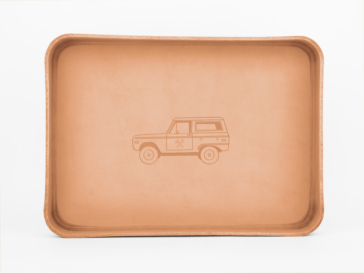A rectangular, tan leather tray with rounded edges features an embossed illustration of a classic SUV on its flat surface. The background is white.