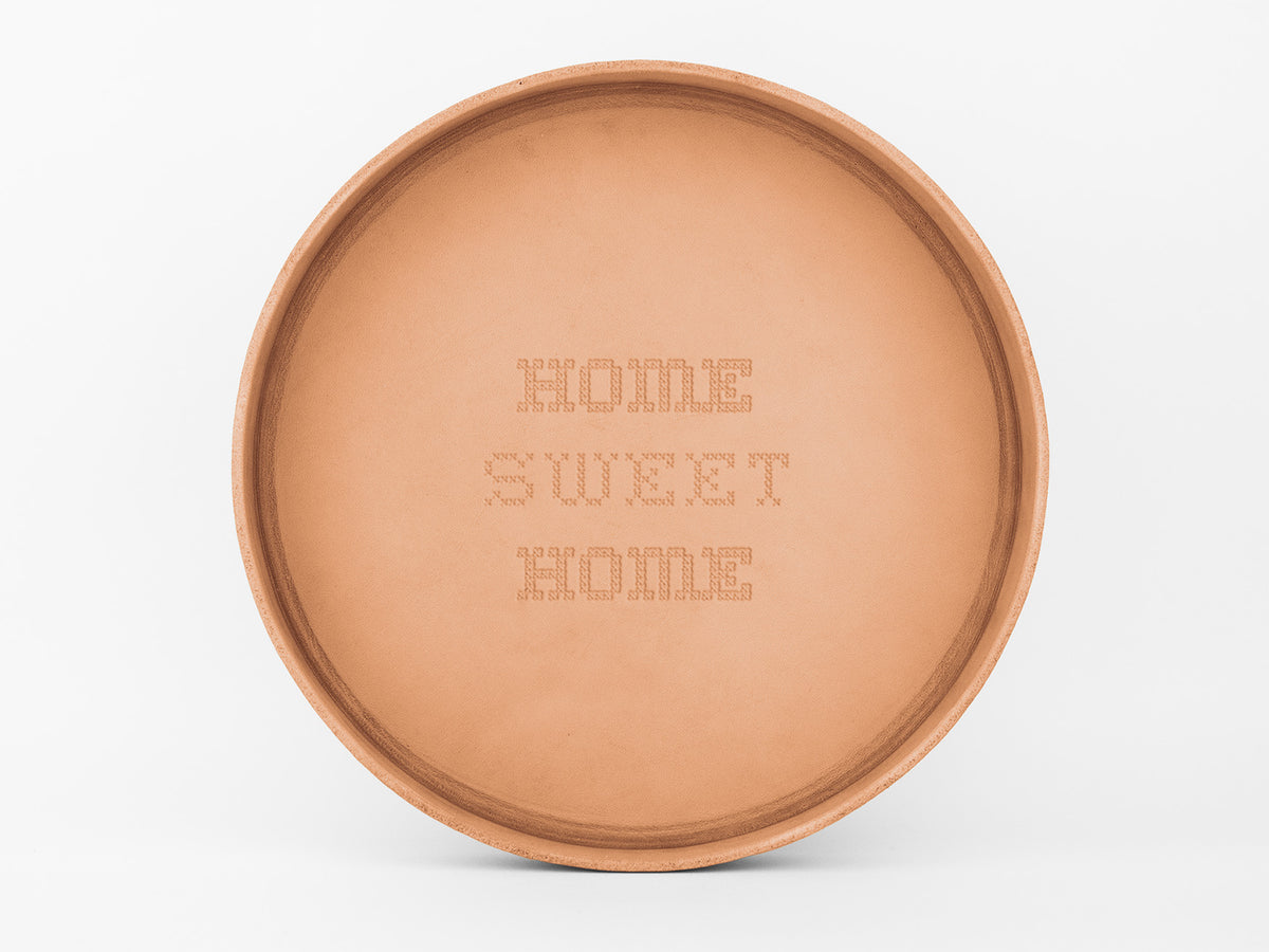 A round, flat, tan-colored tray with the phrase HOME SWEET HOME engraved in a cross-stitch style in the center. The background is plain white.