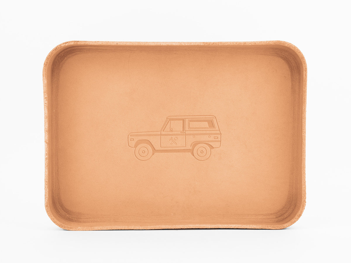 A rectangular, tan leather tray with rounded edges features an embossed outline of a classic SUV with a roof rack in the center. The tray is photographed on a plain white background.