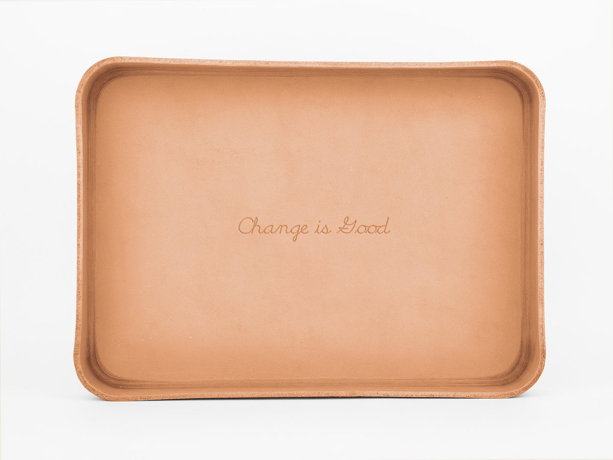 A rectangular, light brown leather tray with rounded corners. The phrase “Change is Good” is engraved in cursive text in the center of the tray. The background is plain white.