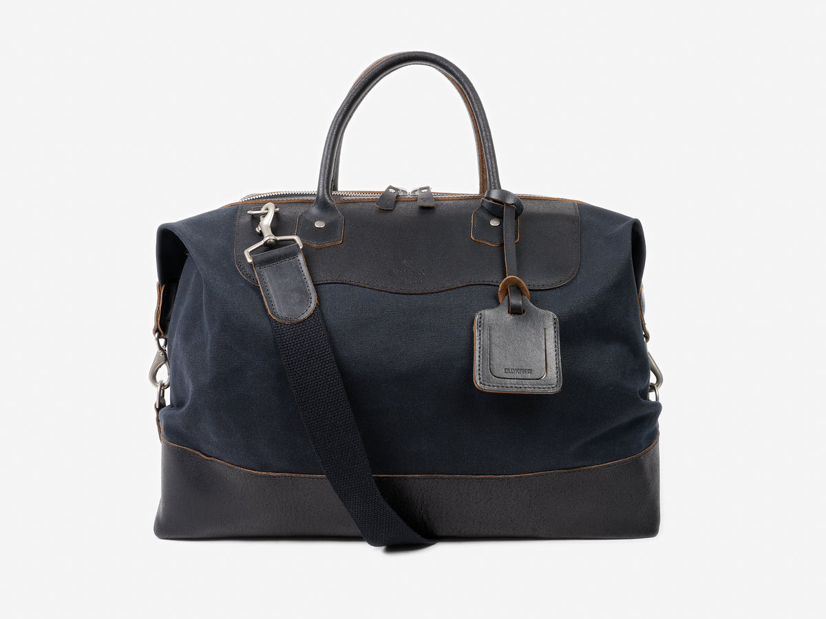 A black and dark navy leather duffel bag with a top zipper, two handles, a detachable shoulder strap, and a leather luggage tag, displayed against a plain light gray background.