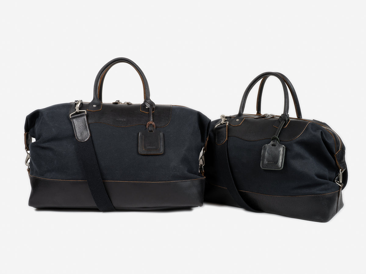 Two black leather and canvas duffel bags with brown stitching, each with handles and an attached luggage tag, are placed side by side on a plain white background.