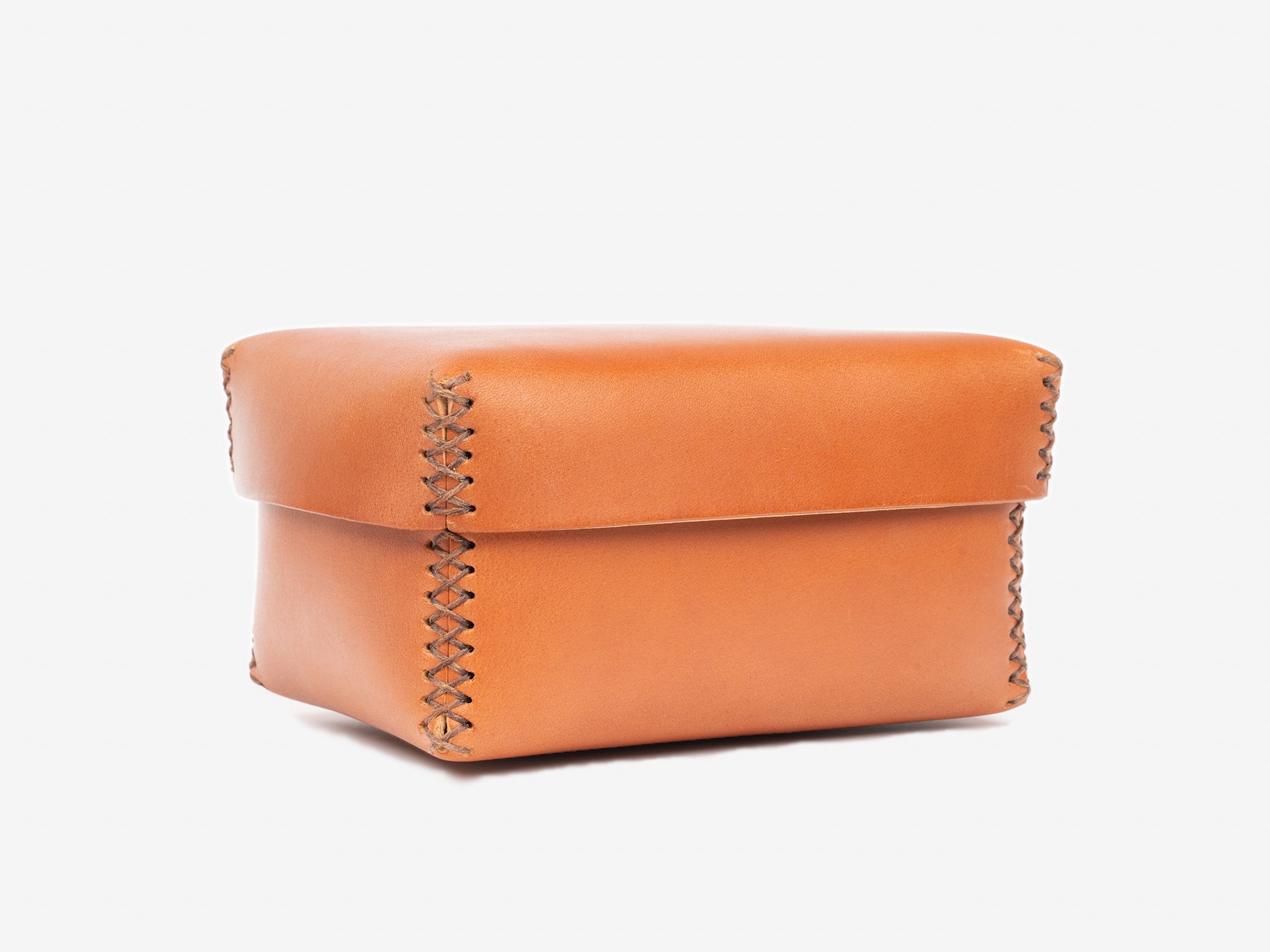 A small, square, tan leather box with visible cross-stitch detailing on the edges and a fitted lid, set against a plain white background.