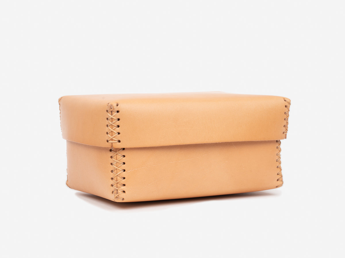 A small, natural leather box with a lid and visible hand-stitched seams along the edges, displayed on a plain white background.