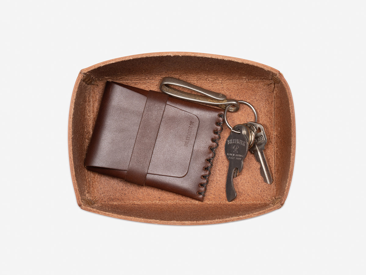A brown card case and a set of keys with a brass keychain rest inside a natural, rectangular open box on a white background.