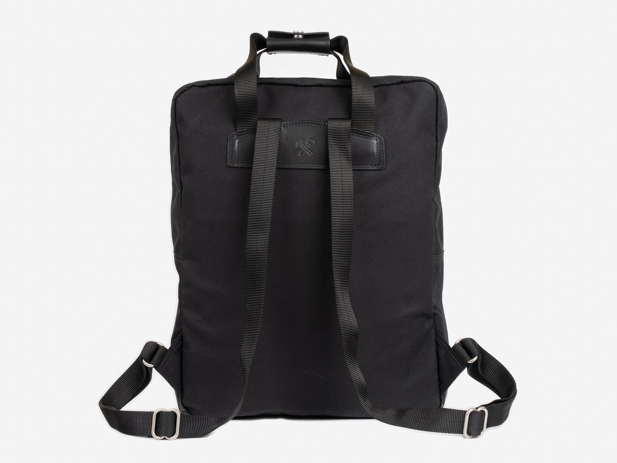 A black waxed canvas backpack shown from the back, featuring adjustable shoulder straps, a top handle, and a rectangular black leather patch near the top center. The background is plain white.