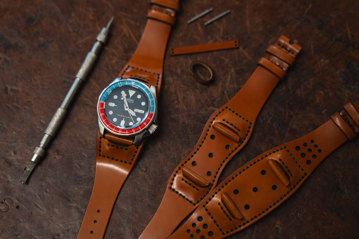 A wristwatch with a blue and red bezel sits on a tan cordovan leather strap, surrounded by additional leather straps, a metal tool, watch pins, and a small leather piece on a rustic wooden surface.