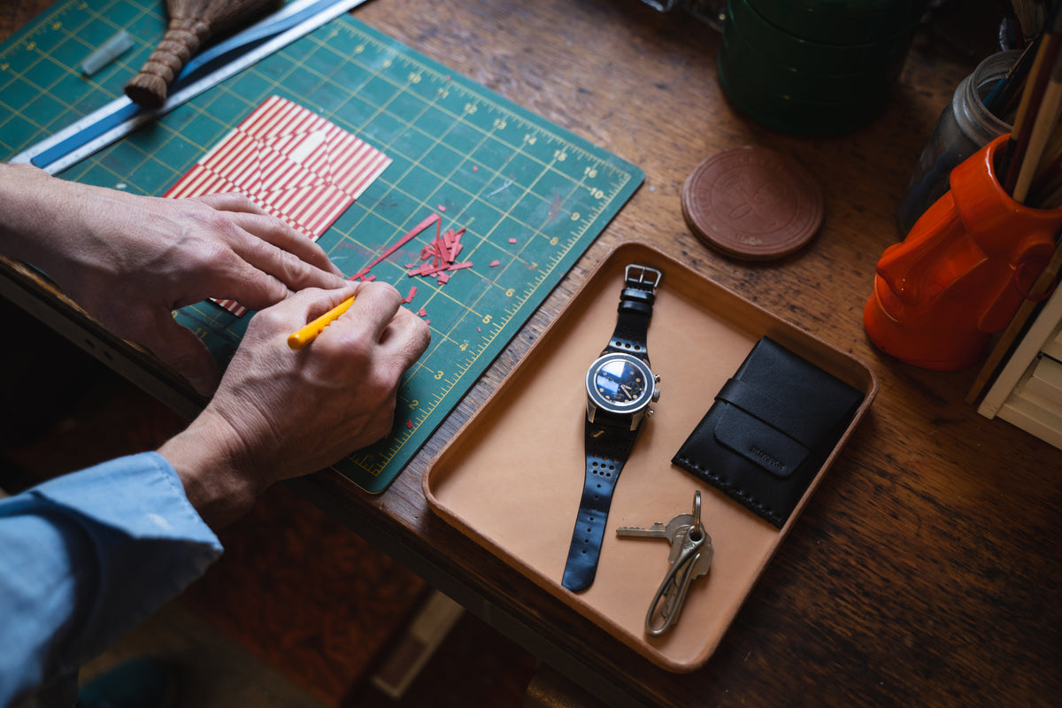 A tray holds a watch with a black cordovan leather strap, a wallet, and keys on a wooden desk. Nearby, a person uses an X-acto knife to cut a red and white striped sheet on a cutting mat.