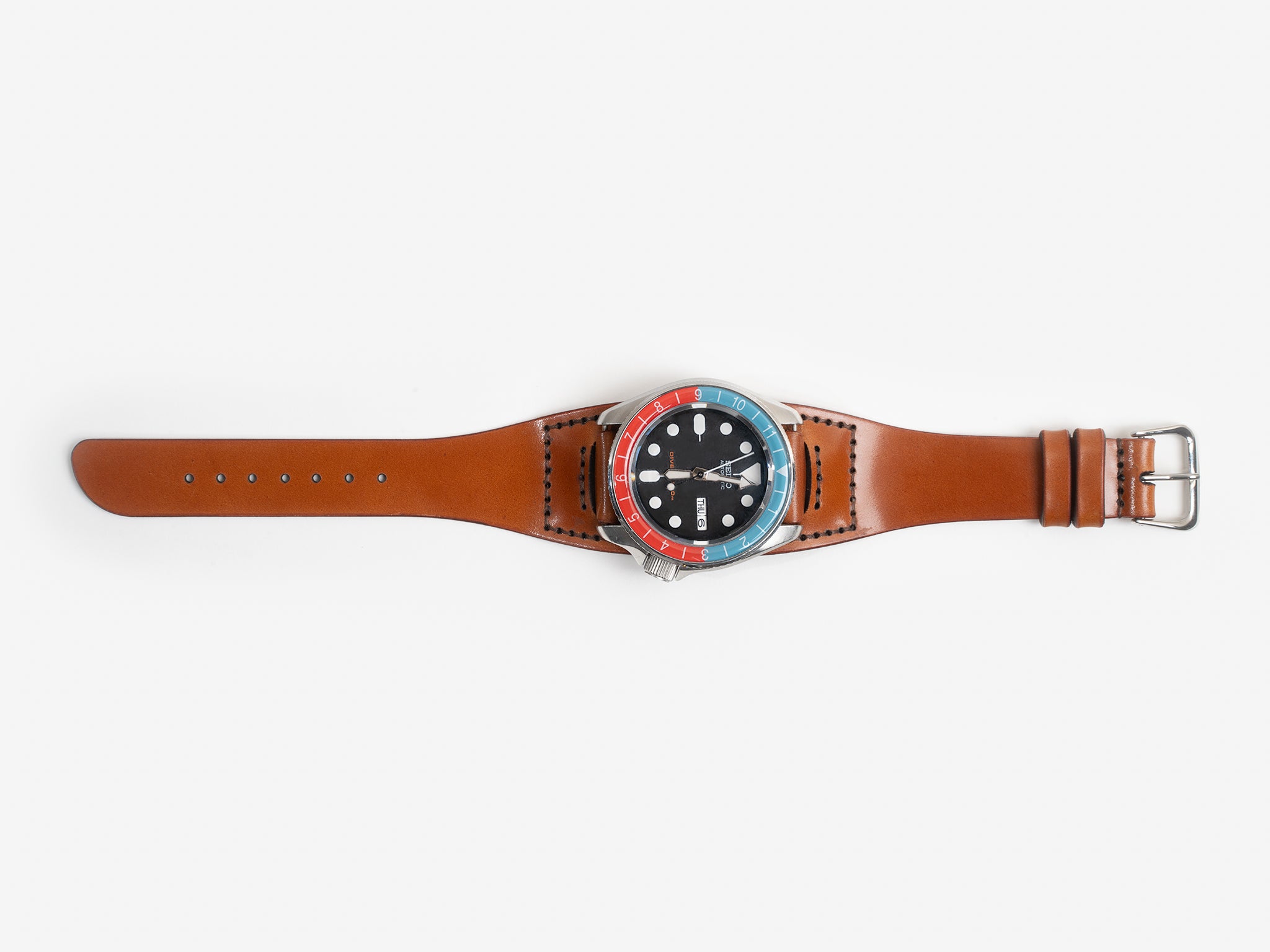 A tan cordovan leather watch strap with a round watch face featuring a blue and red bezel, white hour markers, and silver hands, displayed on a white background.
