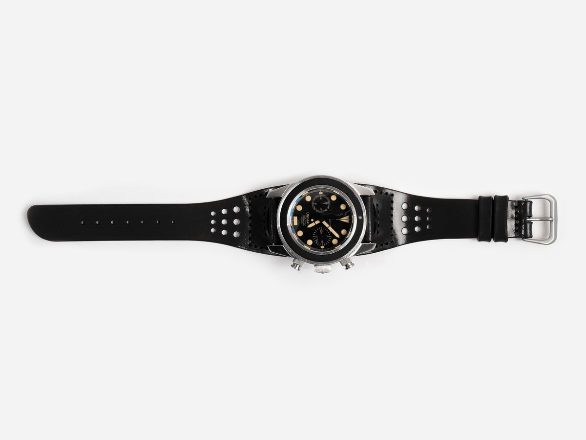 A black cordovan leather watch strap, silver buckle, and a round black dial featuring multiple subdials and gold accents, displayed flat on a white background.