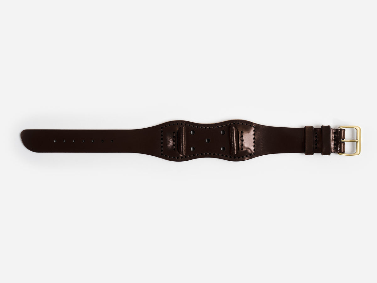 A dark brown cordovan leather watch strap with a brass buckle lies flat on a white background; it has stitched edges and an hourglass shape.