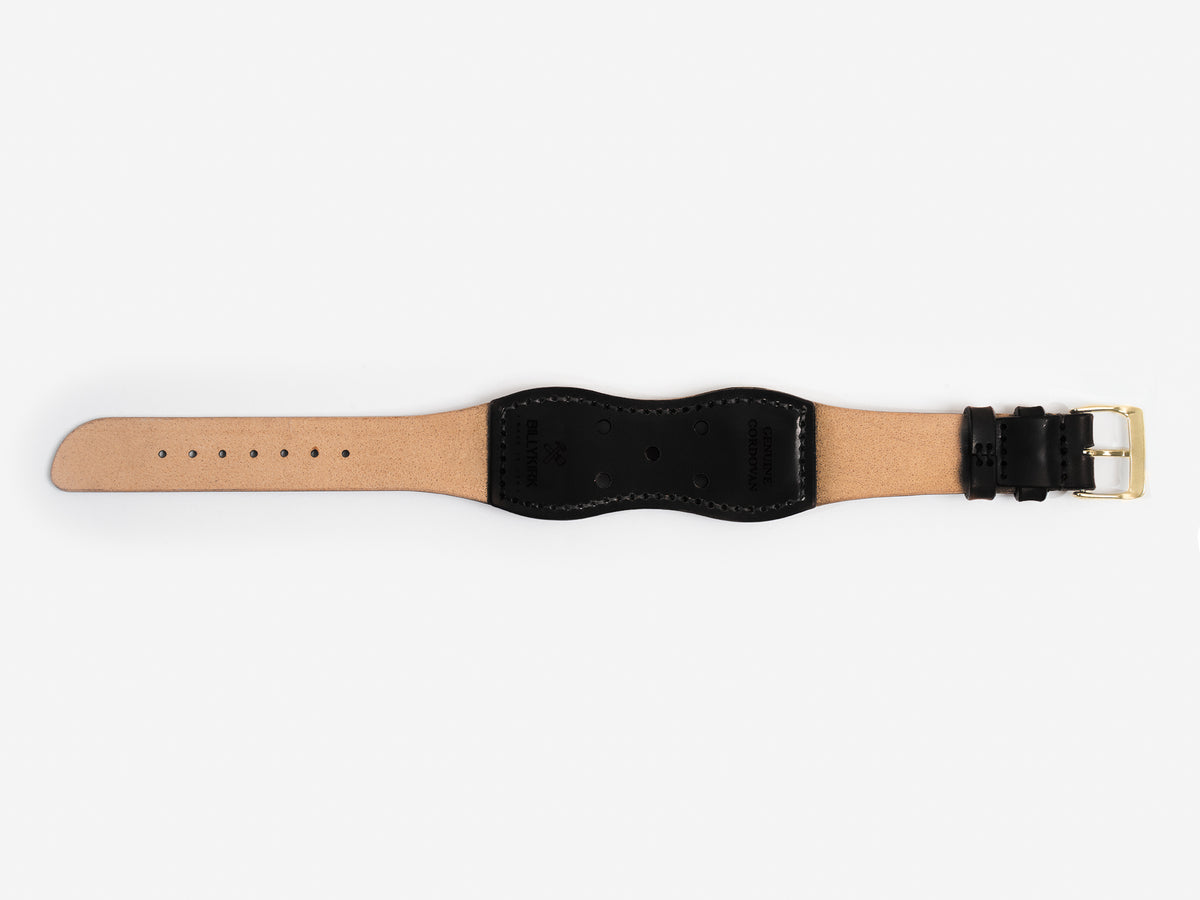 The back of a black cordovan leather watch strap with a brass buckle lies flat on a white background; it has black thread stitched edges and an hourglass shape.