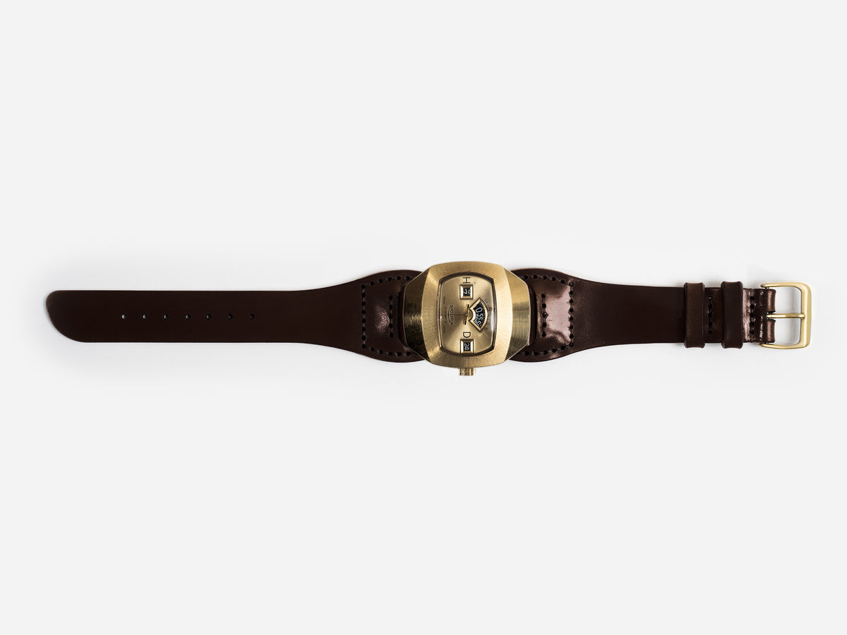 An hourglass-shaped dark brown cordovan leather strap and a large, brass-toned watch face featuring three small rectangular windows displaying the time digitally.