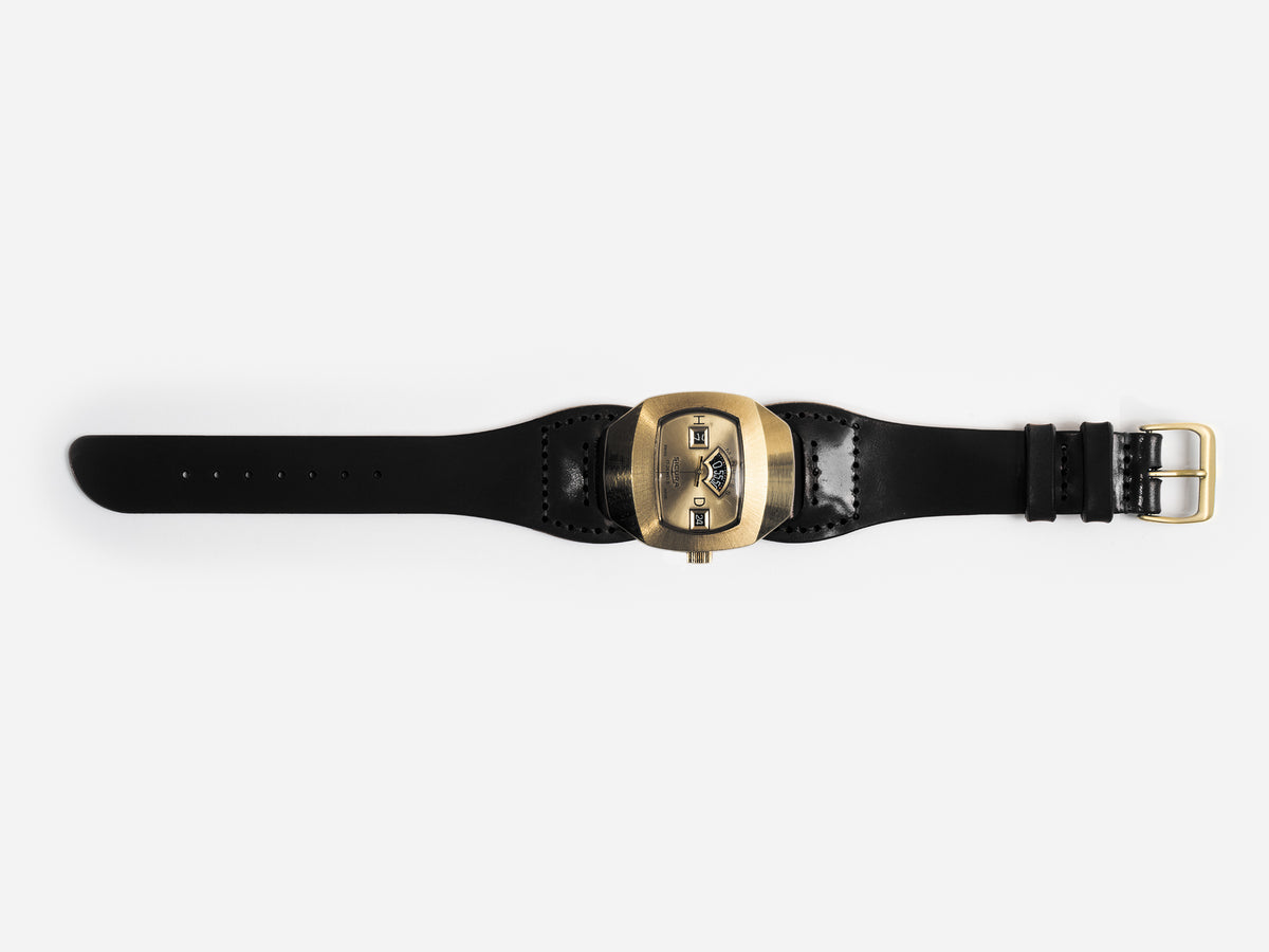 An hourglass-shaped black cordovan leather strap and a large, brass-toned watch face featuring three small rectangular windows displaying the time digitally.