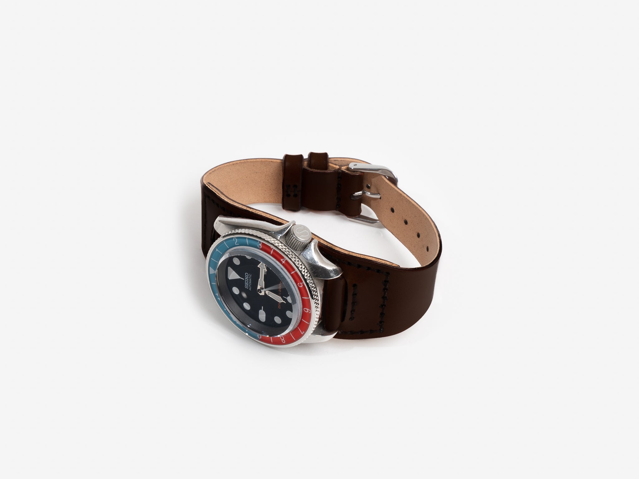 A Seiko watch face with a dark brown cordovan leather strap and a red and blue bezel, displayed on a white background. The watch face features large hour markers and silver hands.
