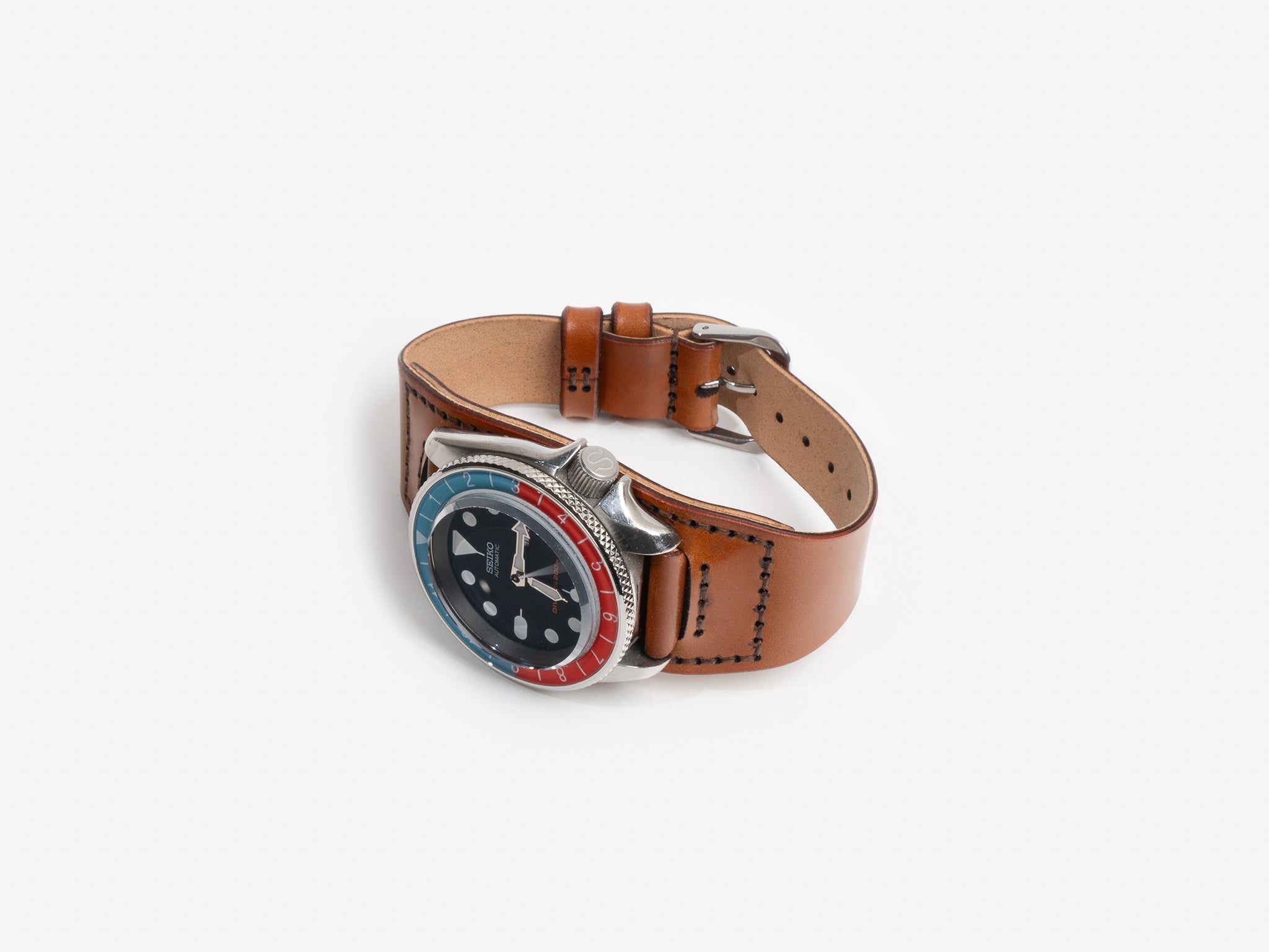 A Seiko watch face with a tan cordovan leather strap and a red and blue bezel, displayed on a white background. The watch face features large hour markers and silver hands.