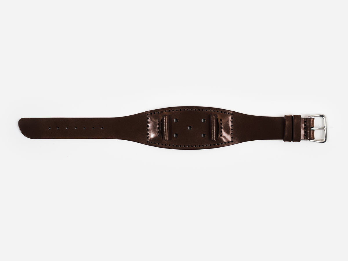 A dark brown cordovan leather watch strap with a silver buckle lies flat on a white background; it has stitched edges and multiple holes for adjustment.
