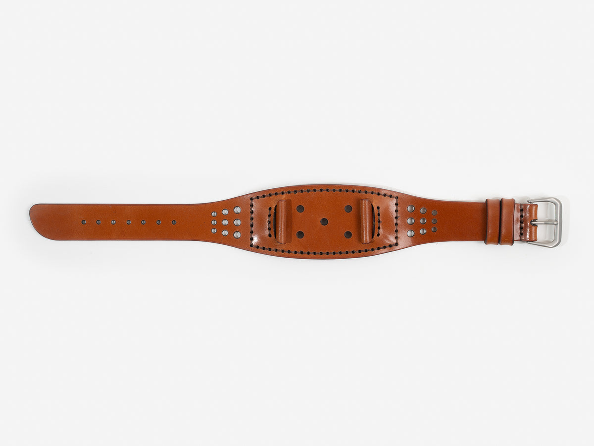 A tan cordovan leather watch strap with a silver buckle lies flat on a white background; it has stitched edges and multiple holes for adjustment.